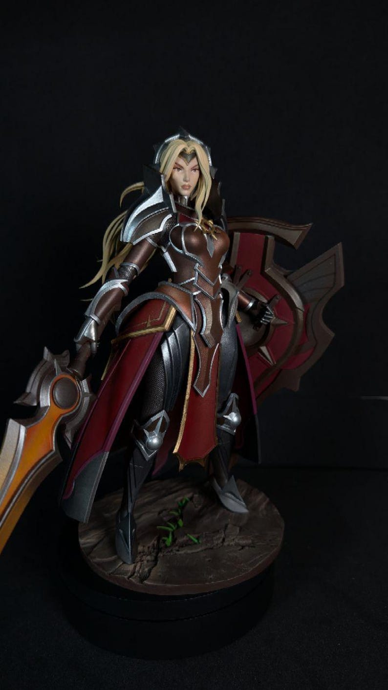 May include: A detailed figurine of a female warrior in armor, holding a sword and shield. The figure is predominantly brown, red, and silver, with blonde hair. The base is a circular platform with a textured, stone-like appearance.