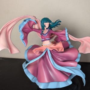 May include: A figurine of a woman in a flowing dress with long sleeves and a scarf. The dress is primarily a deep pink with blue and peach accents. The woman has teal hair and is posed with her arms outstretched.