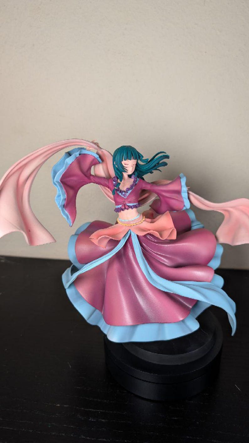 May include: A figurine of a woman in a flowing dress with long sleeves and a scarf. The dress is primarily a deep pink with blue and peach accents. The woman has teal hair and is posed with her arms outstretched.