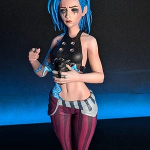 May include: A detailed figurine of a character with blue hair, wearing a black top, striped pants, and brown boots. The figure has blue tattoos and is holding a weapon. The figure is standing on a circular base.