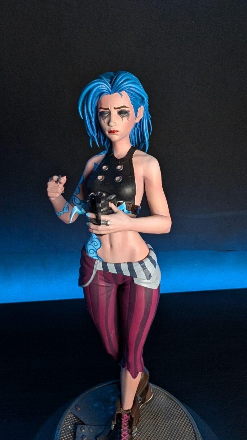 May include: A detailed figurine of a character with blue hair, wearing a black top, striped pants, and brown boots. The figure has blue tattoos and is holding a weapon. The figure is standing on a circular base.