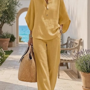 May include: Mustard yellow linen two-piece outfit, comprising a button-down top with a mandarin collar and wide-leg trousers. The outfit is accessorized with a woven tote bag. The background shows a coastal setting with a bench and potted plants.