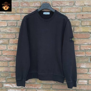 Könnte beinhalten: Ein schwarzes Sweatshirt mit Rundhalsausschnitt und einem kleinen quadratischen Aufnäher auf dem linken Ärmel. Der Aufnäher hat ein gelb-grünes Emblem. Das Sweatshirt hängt an einem schwarzen Kleiderbügel vor einer Backsteinmauer.