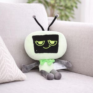 Może przedstawiać: Pluszowa zabawka w kształcie telewizora retro. Ekran jest czarny z uśmiechniętą twarzą i zielonymi oczami. Korpus jest jasnozielony z szarymi ramionami i nogami, i ma dwie czarne anteny z szarymi końc&oacute;wkami.