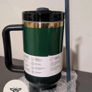 May include: Green travel mug with a black handle and lid, accented with gold trim. Includes a reusable straw. Features double-wall vacuum insulation and a lifetime warranty. The mug is designed for hot and cold beverages.
