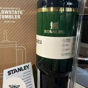 May include: A green and black Stanley Lowstate tumbler with a gold band. The tumbler has the Stanley logo and the text "THE QUENCHER H2.0 LOWSTATE TUMBLER" and "40 OZ | 1.18L". A care and use guide is in front of the tumbler.