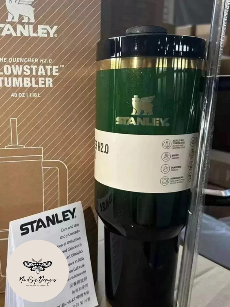 May include: A green and black Stanley Lowstate tumbler with a gold band. The tumbler has the Stanley logo and the text "THE QUENCHER H2.0 LOWSTATE TUMBLER" and "40 OZ | 1.18L". A care and use guide is in front of the tumbler.