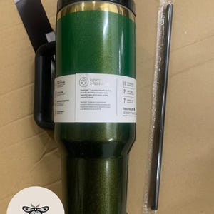 May include: A green glitter travel tumbler with a black handle and lid. The tumbler has a gold band near the top and a white label with text. A black straw is included. The tumbler is designed for hot and cold beverages.