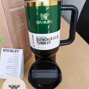 May include: Green and black Stanley Quencher H2.0 tumbler with a black handle and lid. The tumbler has a gold rim and the Stanley logo. The text on the tumbler reads "The Flowstate Quencher H2.0 Tumbler" and "40 oz | 1.18L".