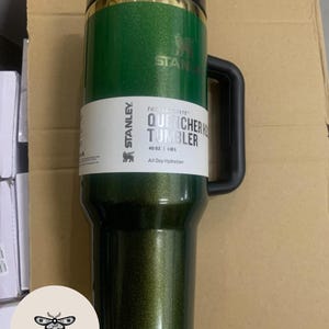 May include: A green Stanley Quencher H2.0 tumbler with a black handle and lid. The tumbler has the Stanley logo and text that reads "QUENCHER TUMBLER 40 OZ | 1.1L All Day Hydration". The tumbler is designed for all-day hydration.
