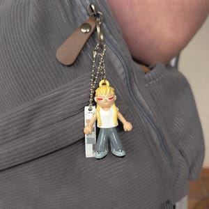 May include: A keychain with a small figurine of a person with blonde hair, red sunglasses, a white top, and grey trousers. The keychain is attached to a grey corduroy backpack with a brown leather zip pull.