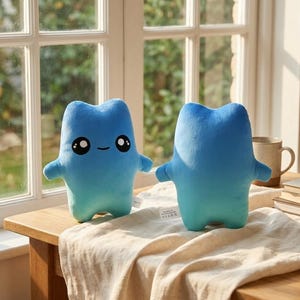 May include: Two plush toys in a soft blue gradient color. One toy features a simple embroidered face with large black eyes and a small smile, while the other is plain blue. Both have a rounded, humanoid shape with short arms and legs.