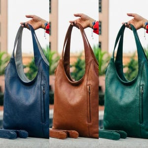 Puede incluir: Tres bolsos de hombro de cuero en azul marino, marrón y verde. Cada bolso tiene una correa larga, un bolsillo con cremallera y una correa a juego. Los bolsos están sostenidos por la mano de una persona.