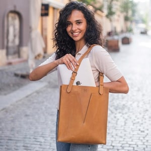 May include: A tan leather tote bag with a laptop inside. The bag has a V-shaped design on the front and adjustable straps. The bag is made of a light brown leather material. The laptop has the Apple logo on the back.