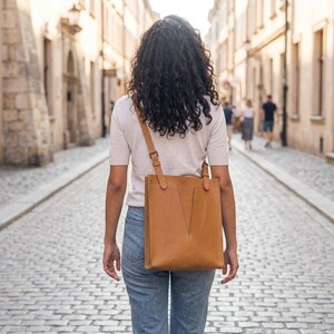 May include: A tan leather tote bag with a shoulder strap. The bag has a rectangular shape with a V-shaped design on the front. The person is wearing a light-colored shirt and blue jeans, walking down a cobblestone street.