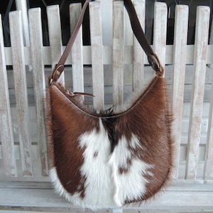 Könnte beinhalten: Eine braun-weiße Schultertasche aus Kuhfell mit einem braunen Lederriemen. Die Tasche hat eine halbmondförmige Form und eine weiche, pelzige Textur. Der Riemen ist mit messingfarbener Hardware befestigt.