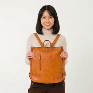 Puede incluir: Una mochila de cuero marrón con un asa superior y correas ajustables para los hombros. La mochila tiene un bolsillo frontal con cremallera y una cremallera para el compartimento principal. El cuero tiene un aspecto natural y texturizado.