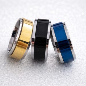 May include: Three rings with a stainless steel band and a colored center band. One ring has a gold center, one is black, and the third is blue. The rings are displayed on a textured, light-colored surface.