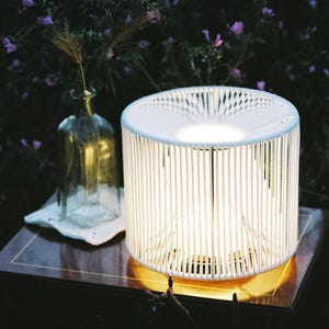 Könnte beinhalten: Eine zylindrische Tischleuchte mit einem weißen Lamellenrahmen, die aus ihrer Mitte ein warmes, leuchtendes Licht ausstrahlt. Die Lampe steht auf einer dunklen, polierten Holzoberfläche neben einer klaren Glasflasche mit getrockneten Blumen.