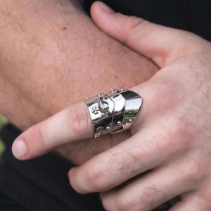 Op de afbeelding: Een zilverkleurige ring met een gesegmenteerd ontwerp, met meerdere platen en klinknagels. De ring wordt aan een vinger gedragen en toont zijn ingewikkelde, pantserachtige uiterlijk. De ring heeft een klein embleem aan de zijkant.