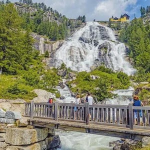 Pode incluir: Uma paisagem pitoresca com uma cachoeira caindo por uma falésia rochosa, cercada por árvores verdes. Uma ponte de madeira com pessoas atravessa um rio. Um edifício é visível na montanha.