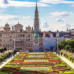 Può includere: Vista panoramica di una città europea con un'alta guglia, edifici e un giardino formale. Il giardino presenta aiuole colorate e siepi curate. Il cielo è blu con nuvole sparse.