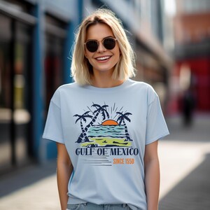 Puede incluir: Una camiseta azul claro con una escena de playa tropical, palmeras, una puesta de sol y el texto "GULF OF MEXICO SINCE 1550" en blanco y naranja. La camiseta la lleva una persona con gafas de sol.