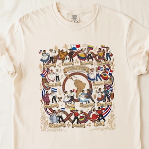 May include: Cream-colored t-shirt featuring a detailed illustration of Latin American dancers and flags. The design includes the text "Tradicional Latinoamerica El Ritmo de la Tierra Las Danzas y Sus Banderas" and country names like Mexico and Argentina.