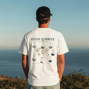 May include: A white t-shirt featuring a dark green graphic on the back. The design displays a world map with seven circular illustrations of mountain peaks labelled Seven Summits, including Everest, Aconcagua, Denali, Kilimanjaro, Vinson, Puncak Jaya, and Elbrus.