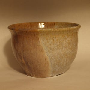 May include: A handmade ceramic bowl with a rounded shape and a wide opening. The bowl features a two-tone glaze, with a warm brown color on one side and a lighter, off-white hue on the other. The rim is slightly flared.