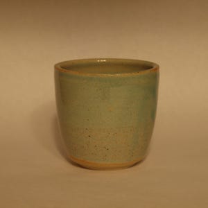 May include: A handmade ceramic cup with a rounded shape and a speckled, light teal glaze. The rim and base of the cup have a natural, unglazed appearance. The cup is approximately 9 cm tall and 7.6 cm wide.