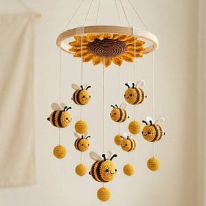 Op de afbeelding: Een hangende mobiel met een grote gehaakte zonnebloem bevestigd aan een rond houten frame. Verschillende gehaakte bijen met gele en zwarte strepen en witte vleugels, samen met kleine gele bolletjes, hangen aan witte koordjes aan de zonnebloem.