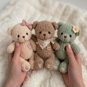 以下が含まれることがあります： クリーム色、茶色、セージグリーンの3体の小さなかぎ針編みのぬいぐるみテディベア。クリーム色のベアにはピンクのリボンとハートのパッチが付いています。茶色のベアは花柄のバンダナと小さなクマのボタンを身に着けています。緑色のベアには花のアクセサリーが付いています。