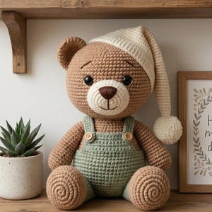 May include: A crocheted teddy bear with light brown yarn, wearing a cream-colored nightcap with a pom-pom and sage green overalls with two small wooden buttons. The bear has black plastic eyes, a dark brown stitched nose, and a cream-colored muzzle.