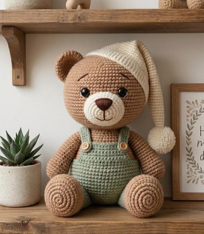 May include: A crocheted teddy bear with light brown yarn, wearing a cream-colored nightcap with a pom-pom and sage green overalls with two small wooden buttons. The bear has black plastic eyes, a dark brown stitched nose, and a cream-colored muzzle.