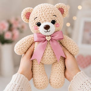 Puede incluir: Un oso de peluche tejido a ganchillo en color beige con hocico blanco, nariz marrón oscuro y ojos negros con pestañas largas. El oso tiene detalles rosados en el interior de las orejas y mejillas, decorado con un lazo de satén rosa y un botón.