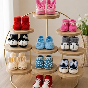 May include: A collection of various crocheted baby booties displayed on a tiered gold metal stand. The booties feature diverse designs including bunny ears, bows, stars, and animal faces in colors such as pink, red, blue, black, gray, and cream.