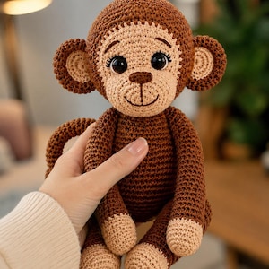May include: A brown and beige crocheted monkey toy featuring a round head, large ears, dark plastic eyes, and a stitched smile. The body is crafted with dark brown yarn, while the face, ears, and feet are accented with a lighter beige yarn colour.
