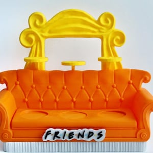 May include: A miniature orange sofa with a yellow decorative top, resembling the Central Perk couch from the TV show "Friends." The sofa has a white base and the word "FRIENDS" is displayed on the front.