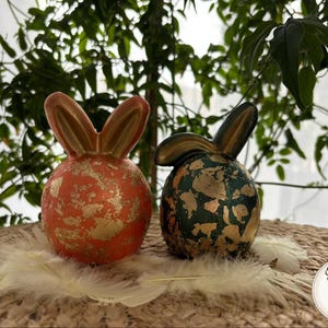 May include: Two ceramic bunny ornaments. One is orange with gold leaf detailing, the other is dark green with gold leaf. Both have upright ears and are displayed on a woven surface with feathers.