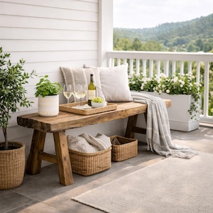 Op de afbeelding: Een rustieke houten bank met een dienblad met wijn en glazen, kussens en een grijze deken. Rotan manden en een potplant staan op het terras. Een witte plantenbak met bloemen staat op de reling.