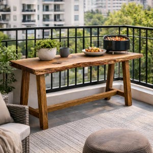 Puede incluir: Mesa de exterior de madera rústica con borde natural. La mesa tiene patas en ángulo y está en un balcón. Una pequeña planta en maceta, una taza, un plato de comida y una pequeña parrilla están sobre la mesa. La mesa mide unos 150 cm de largo.
