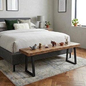 Puede incluir: Un banco rústico de madera con patas de metal negro, colocado al pie de una cama. El banco está adornado con objetos decorativos, como un pájaro de cerámica, un búho y un elefante, junto con pequeños cuencos y jarrones. La cama tiene un marco tapizado gris y está cubierta con ropa de cama blanca.