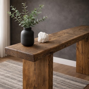 May include: A rustic wooden bench with a dark finish. A black vase with green foliage and a white stone sit on the bench. The bench has a thick, rectangular top and sturdy legs, showcasing natural wood grain.