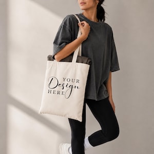 Può includere: Borsa tote color crema con la scritta "YOUR Design HERE" in corsivo nero. La borsa ha una forma rettangolare e manici lunghi. La borsa viene portata a spalla. La borsa è in tessuto chiaro.