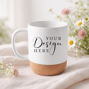 Puede incluir: Taza de cerámica blanca con base de corcho, con el texto "YOUR Design HERE" en escritura negra. La taza tiene un asa curva y está sobre un fondo suave y neutro con flores.