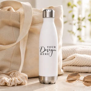 Op de afbeelding: Witte geïsoleerde waterfles met een zilveren dop en bodem. De fles heeft de tekst "YOUR Design HERE" in zwart script. Een beige draagtas, scrunchie, zonnebril en gevouwen trui staan op de achtergrond.