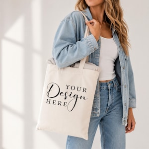 Könnte beinhalten: Cremefarbene Tragetasche mit dem Text "YOUR Design HERE" in schwarzer Schrift. Die Tasche wird von einer Person getragen, die eine hellblaue Jeansjacke und Jeans trägt. Die Tasche hat ein schlichtes, minimalistisches Design.