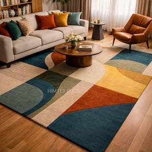 May include: A modern geometric rug in a living room setting. The rug features a pattern of blue, yellow, orange, and beige shapes. The room includes a sofa, armchair, and a wooden coffee table. The rug's dimensions are in centimeters.