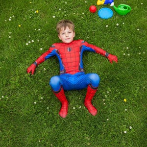 Puede incluir: Un ni&ntilde;o con un disfraz de Spider-Man rojo y azul yace sobre hierba verde. El disfraz presenta un emblema de ara&ntilde;a y patrones de telara&ntilde;a. Peque&ntilde;os juguetes est&aacute;n dispersos cerca, incluyendo una pelota roja y una regadera verde.